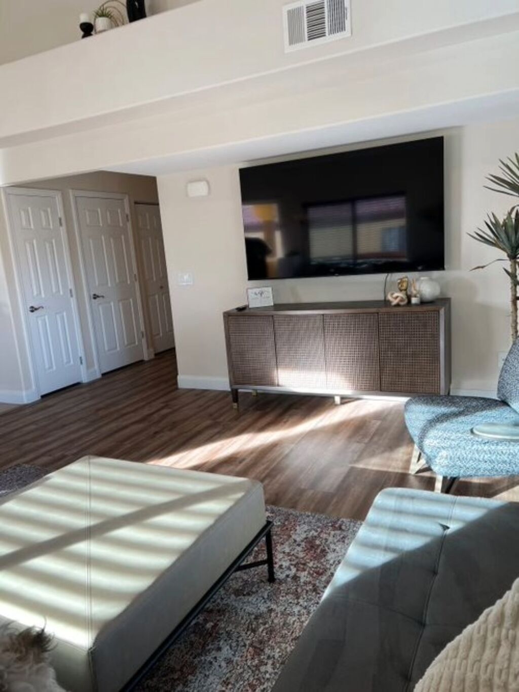 A living room with a flat-screen TV, a grey sofa, a patterned rug, and a wooden floor.