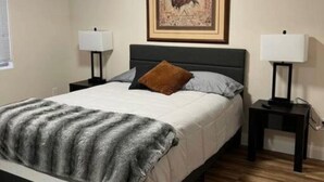Une chambre à coucher comprenant un lit, deux tables de chevet, une commode, un tableau encadré au mur et une fenêtre avec des stores.