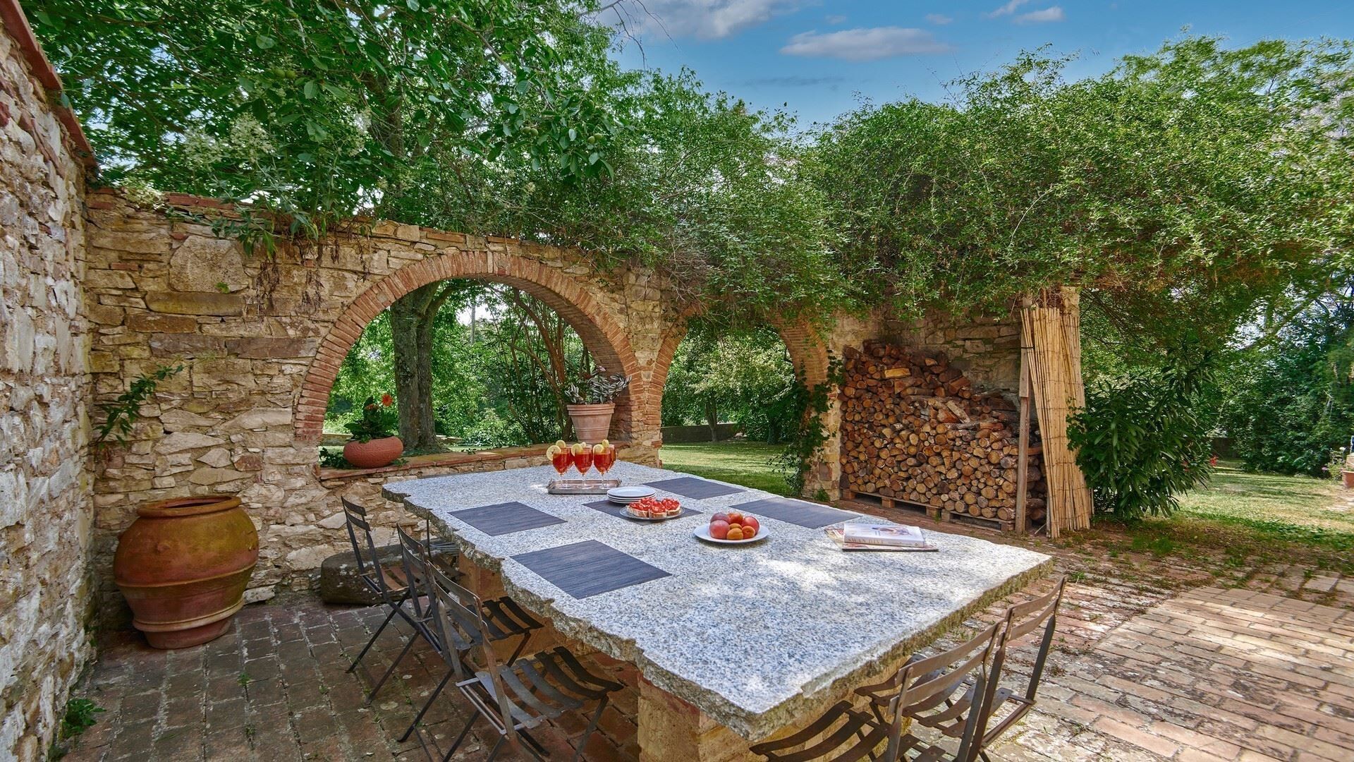 Un'area pranzo all'aperto con tavolo e sedie in pietra, circondata da un muro di pietra e alberi.
