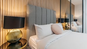 A modern hotel room with a large bed, a headboard with vertical wooden slats, a round gold lamp, and a mirror reflecting the room. - Grace At Mosman - Service Apartments (Mosman)