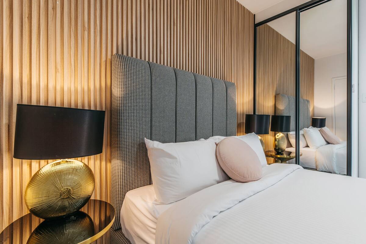 A modern hotel room with a large bed, a headboard with vertical wooden slats, a round gold lamp, and a mirror reflecting the room.