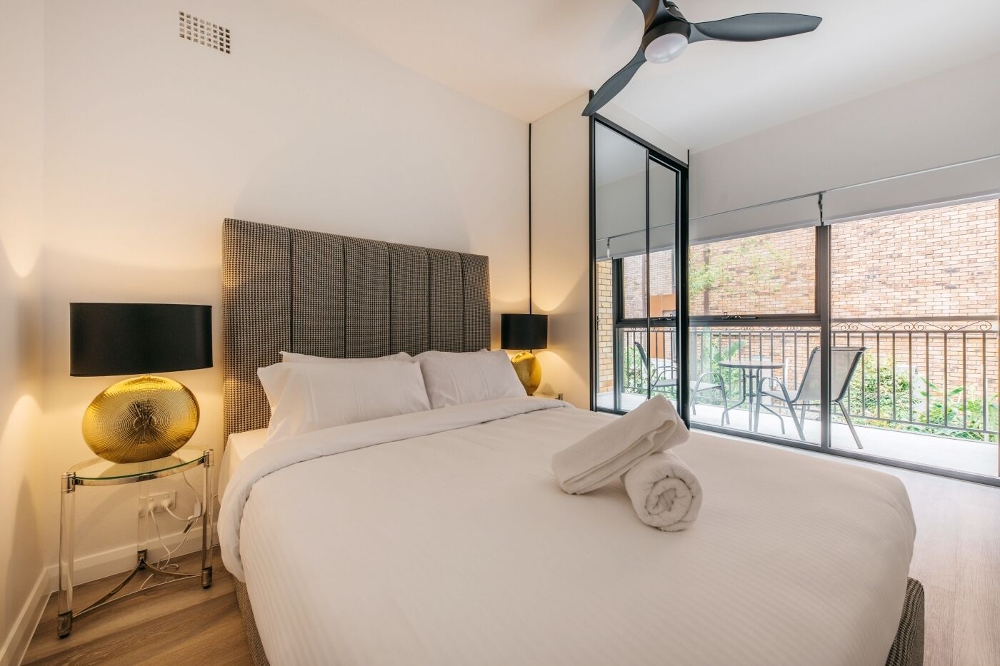 A modern hotel room with a large bed, two bedside tables with lamps, a ceiling fan, and a balcony with outdoor seating.