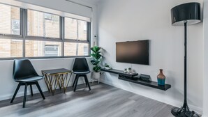 A modern living room with a large window, two black chairs, a small table, a TV mounted on the wall, a floor lamp, and a shelf with decorative items. - Grace At Mosman - Service Apartments (Mosman)