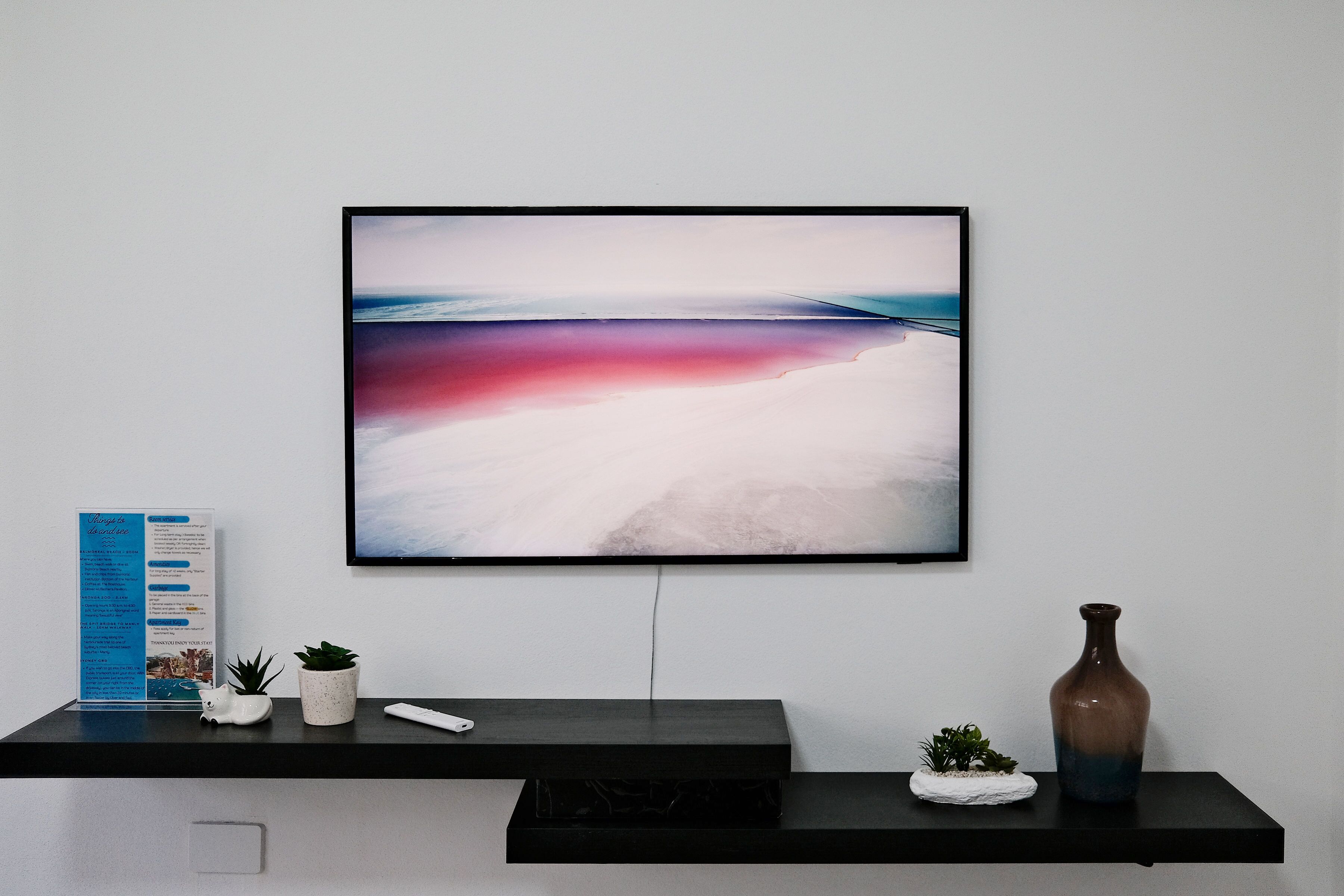 A framed artwork on a wall, a small potted plant, a remote control, and a vase on a black shelf.
