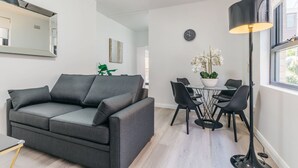 A modern living room with a grey sofa, a dining table with chairs, a black floor lamp, and a large mirror on the wall. - Grace At Mosman - Service Apartments (Mosman)