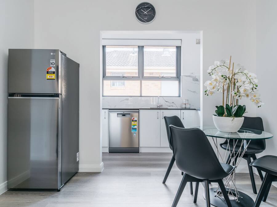 A modern kitchen with a stainless steel refrigerator, a dishwasher, and a dining area with a glass table and black chairs.