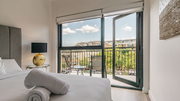 A hotel room with a large bed, a round table with chairs, a balcony with a view of buildings and greenery, and a wall-mounted lamp. - Grace At Mosman - Service Apartments (Mosman)