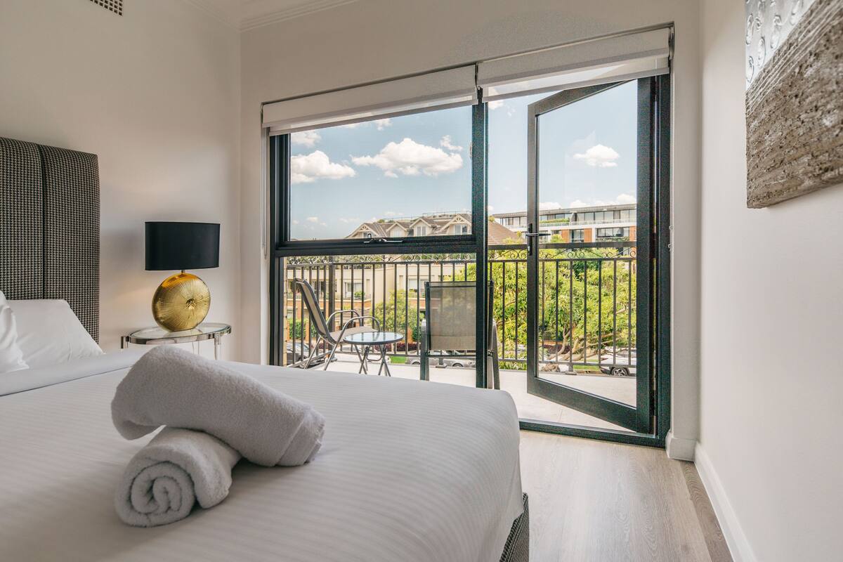 A hotel room with a large bed, a round table with chairs, a balcony with a view of buildings and greenery, and a wall-mounted lamp.