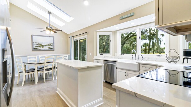 Una cocina moderna con una isla central, armarios blancos y grandes ventanales que ofrecen vistas a árboles.