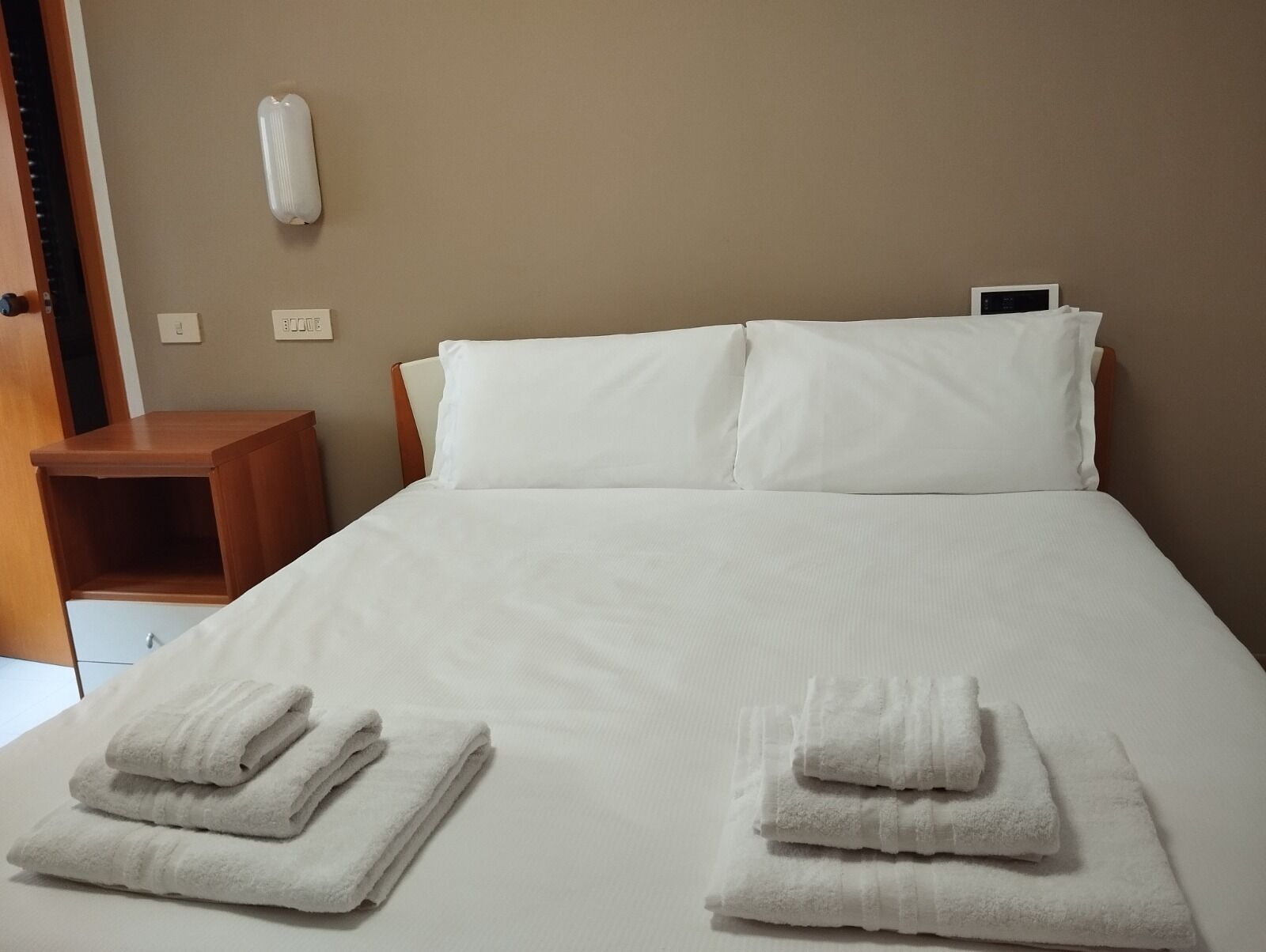 Ein ordentlich bezogenes Bett mit weißen Leinen und Kissen, ein Nachttisch aus Holz mit einer Lampe und ein an der Wand befestigtes Thermostat.