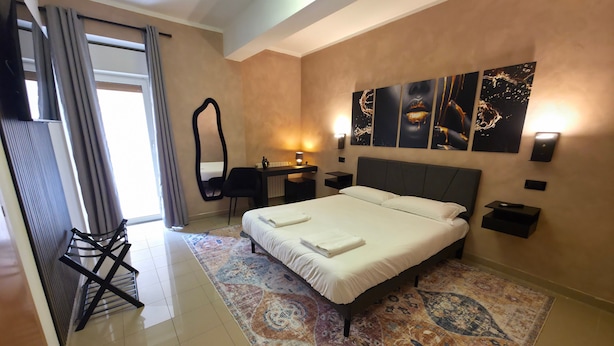 A hotel room with a bed, a desk, a chair, a mirror, and artwork on the wall. - AH Roma (Rome)