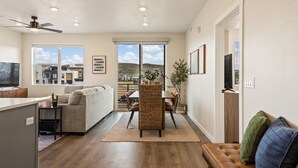 Ett modernt vardagsrum med en soffa, ett matbord och stolar, en takfläkt och utsikt över en stadssiluett genom stora fönster.