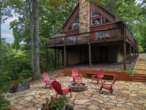 Una cabaña con chimenea de piedra, sillas Adirondack rojas y una terraza con vista a un bosque.