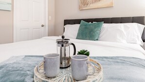 A neatly made bed with white linens, a gray headboard, and a teal pillow. A coffee maker and two gray mugs are on a woven tray beside the bed. - Fast WiFi, King Bed, Gym w/ Free Parking (Huntsville)