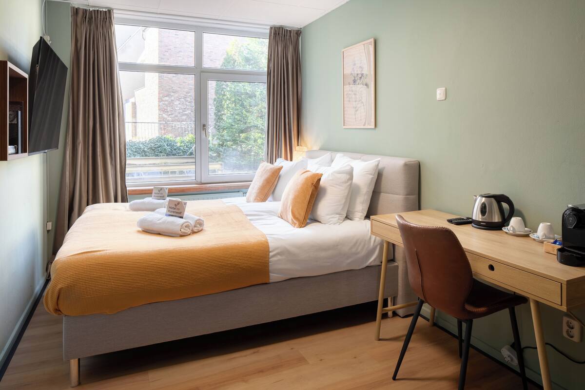 Une chambre d’hôtel équipée d’un lit, d’un bureau, d’une chaise, d’une fenêtre avec des rideaux, d’un téléviseur à écran plat et d’un tableau encadré au mur.