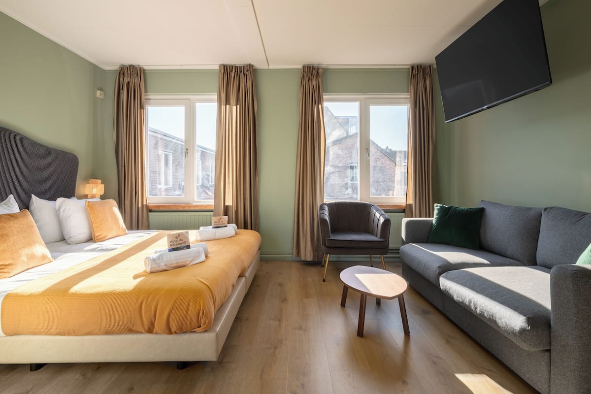 Une chambre d’hôtel avec un lit, un canapé, une chaise, une télévision et une fenêtre avec des rideaux.
