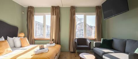 A hotel room with a bed, a sofa, a chair, a TV, and a window with curtains.