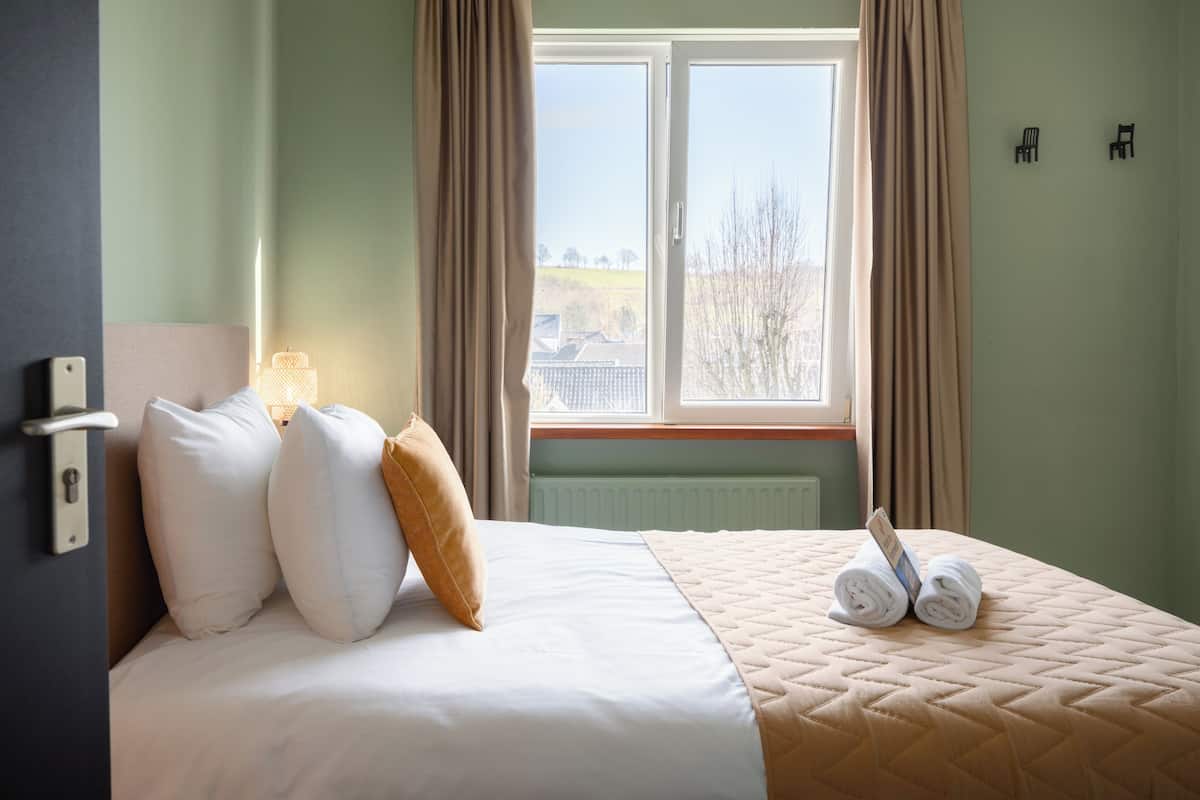 Une chambre d’hôtel avec un lit, des oreillers, une fenêtre avec des rideaux, un radiateur et un luminaire fixé au mur.