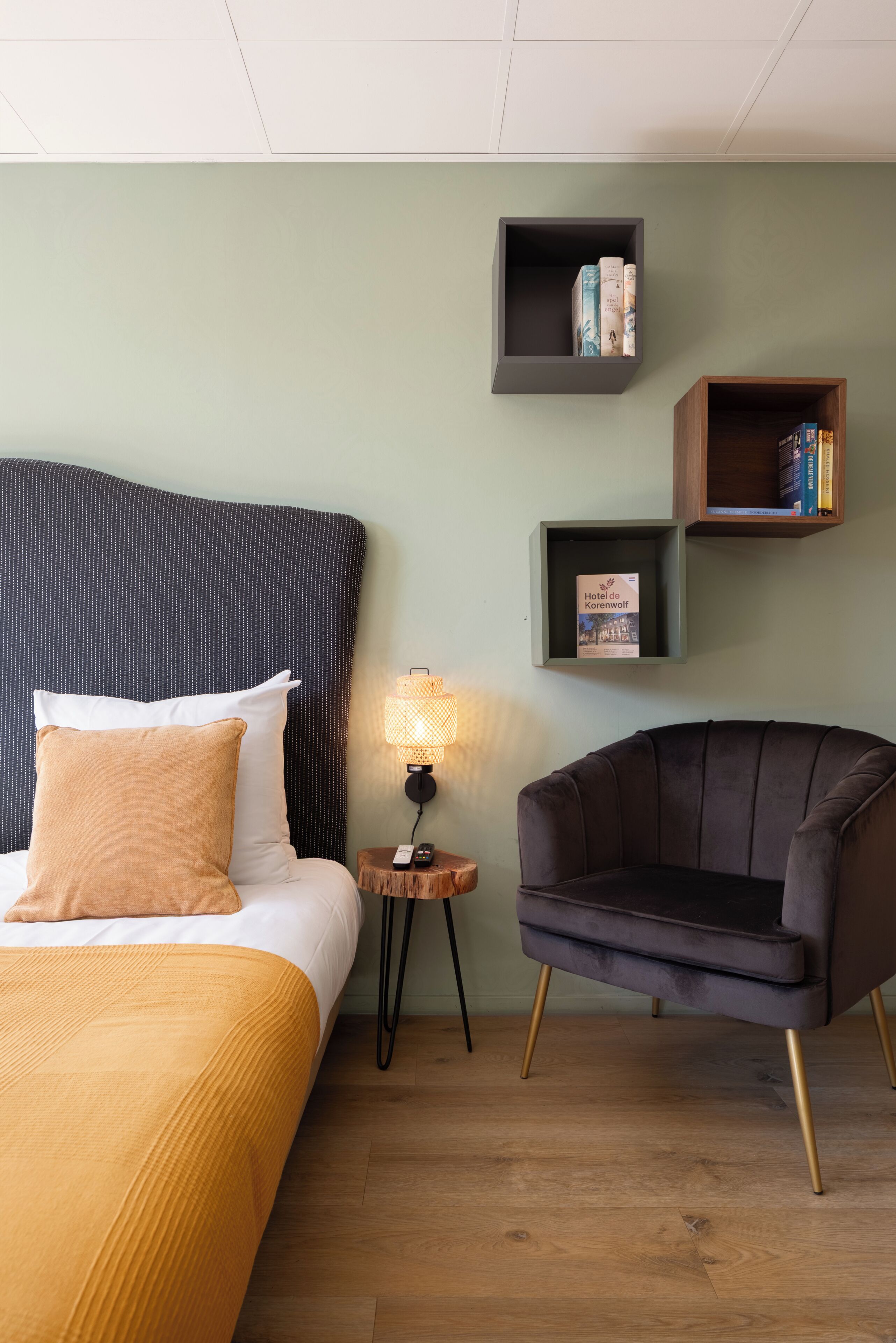 Habitación de hotel con cama, una silla, mesita de noche con lámpara y dos estantes con libros y un folleto.