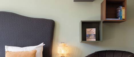 A hotel room with a bed, a chair, a nightstand with a lamp, and two shelves with books and a brochure.