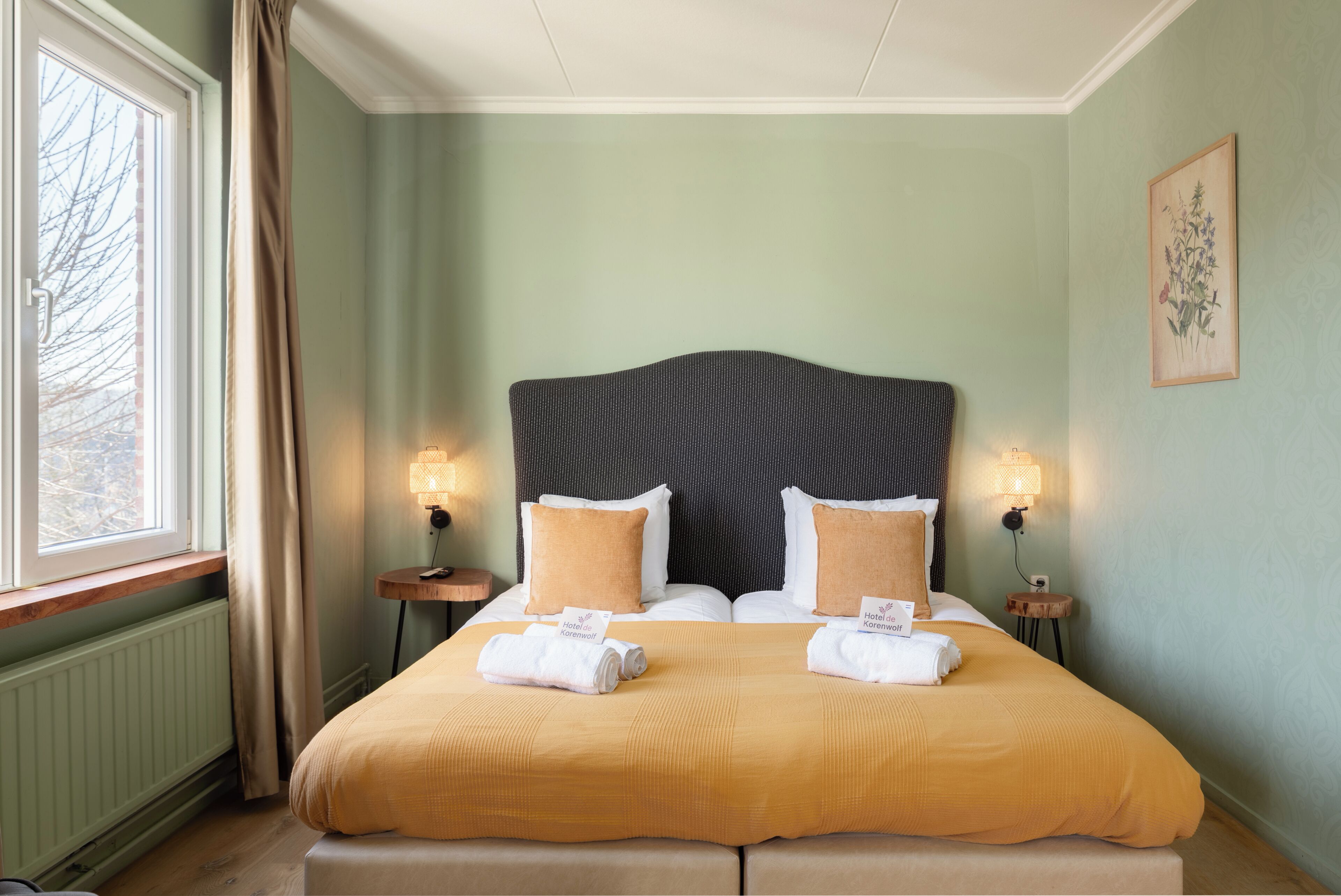 A double bed with a headboard, two bedside tables with lamps, a window with curtains, and a framed botanical print on the wall.
