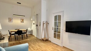 Amplia sala de estar con piso de madera, un sofá azul, una zona de comedor con mesa y sillas, un televisor montado en la pared y una lámpara colgante.