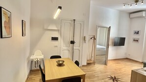 Una habitación moderna y bien iluminada con una mesa de comedor de madera, sillas negras y una pequeña mesa blanca. Hay una puerta blanca, un perchero y un televisor montado en la pared.