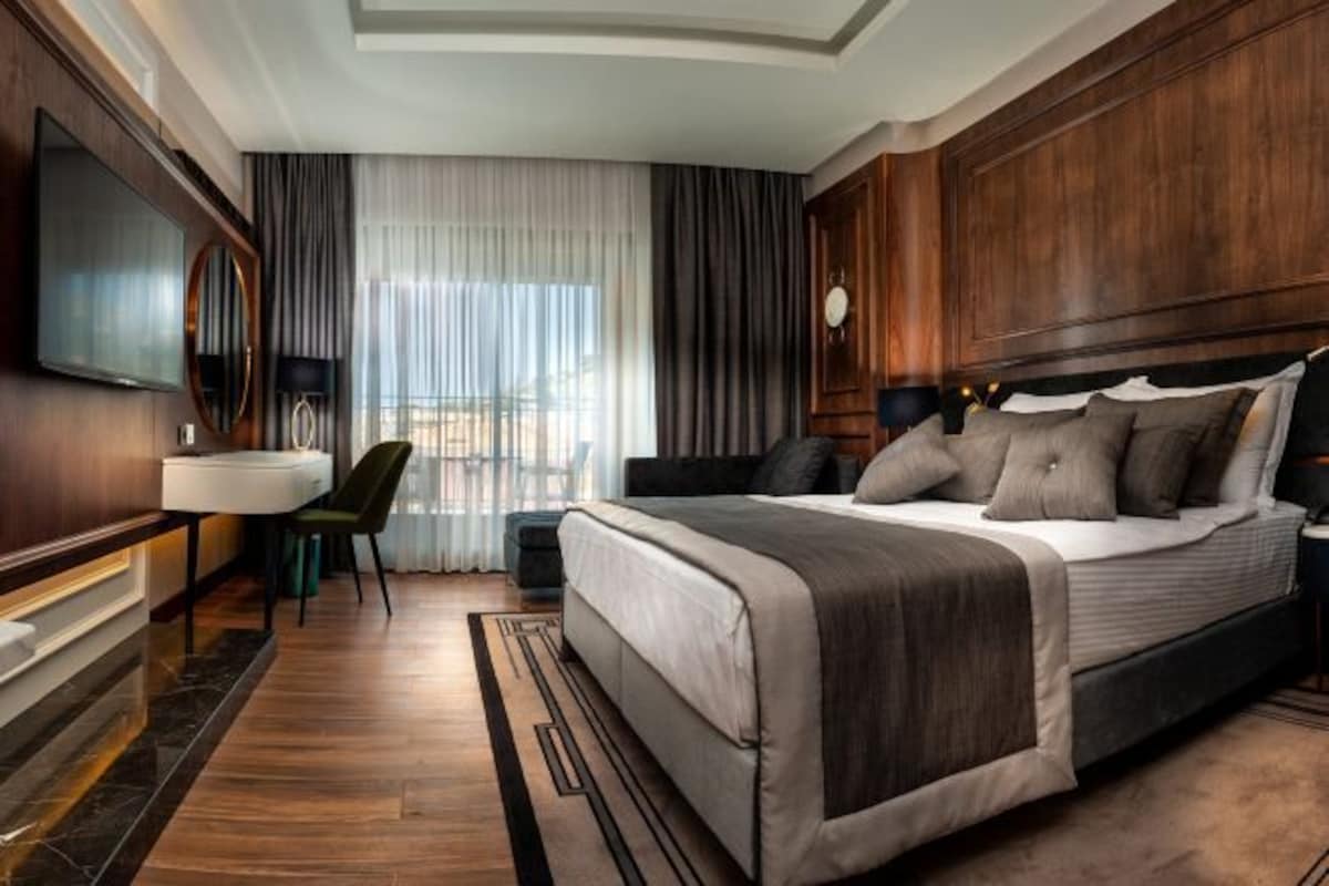 Habitación de hotel moderna con una cama grande, un escritorio con una silla, televisión y vistas al exterior a través de ventanas amplias.