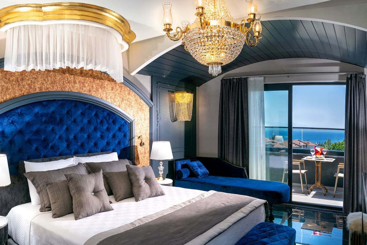 A luxurious bedroom with a tufted headboard, a large bed with multiple pillows, a nightstand with a lamp, a blue sofa, and a balcony with a table and chairs.