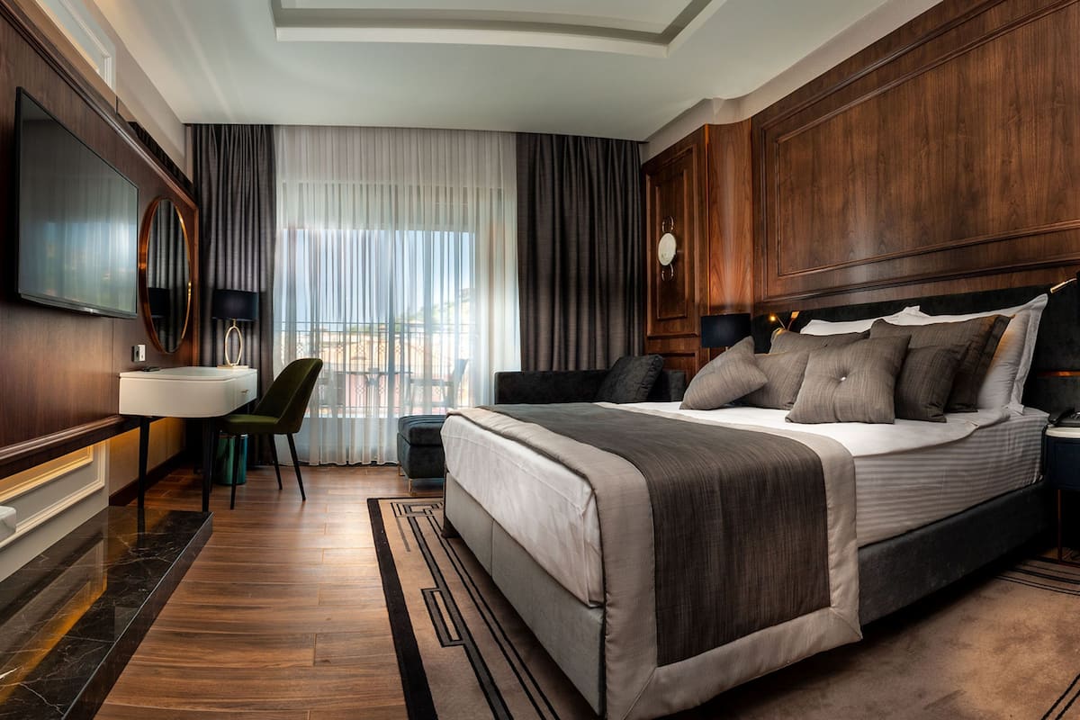A hotel room with a large bed, a desk with a chair, a vanity, and a television.