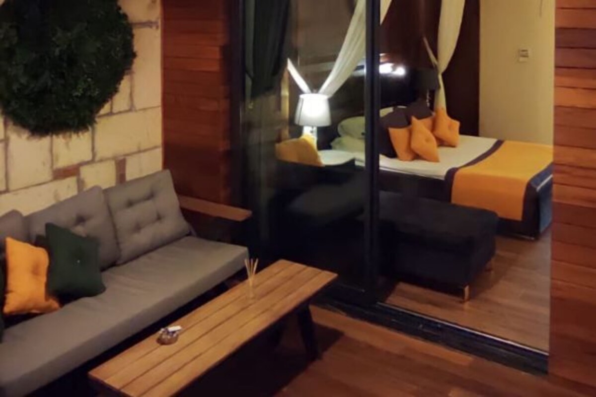 A modern room with a grey sofa, a wooden coffee table, a glass partition, and a bed with orange bedding.