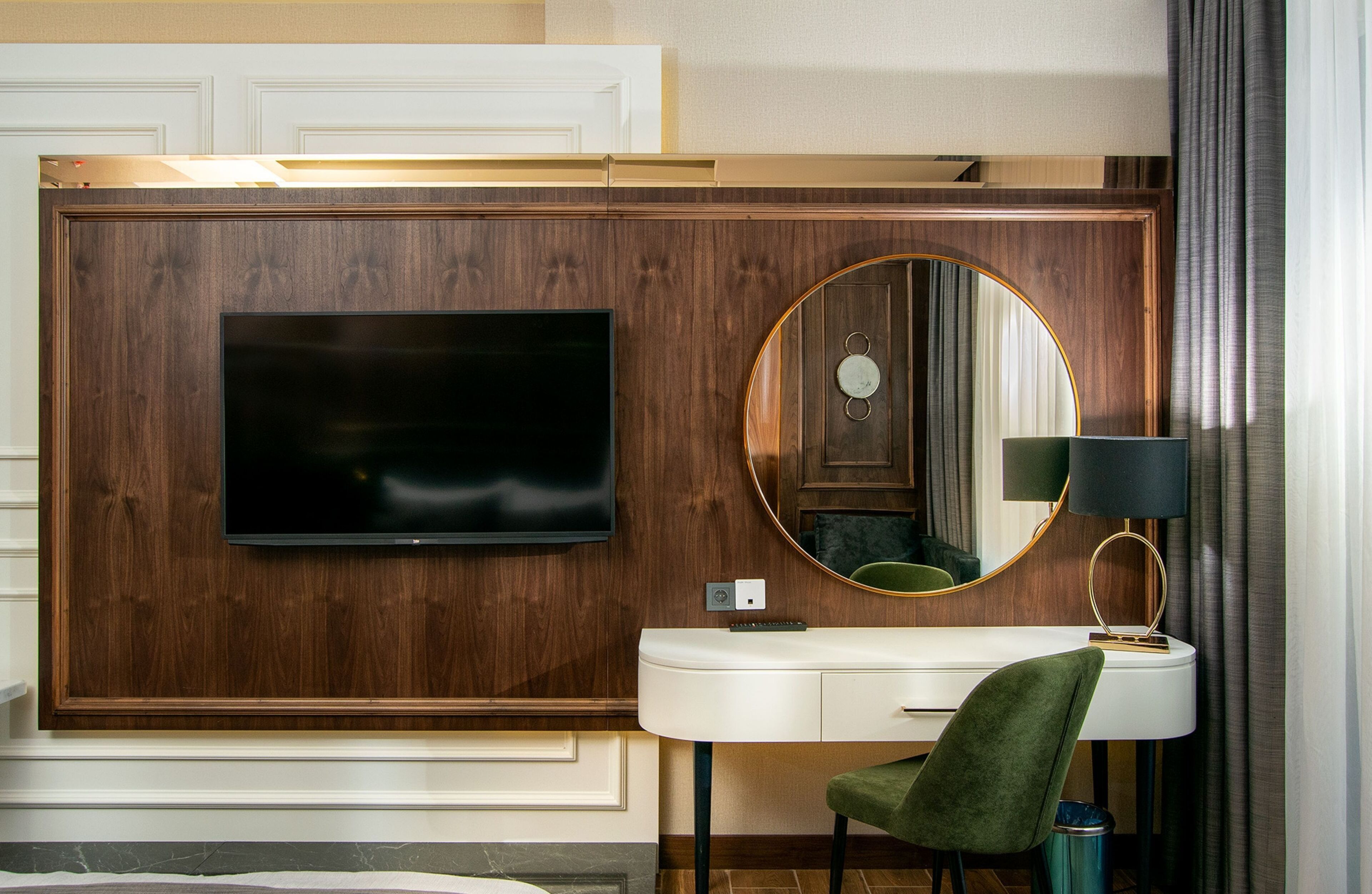 Habitación de hotel con una televisión de pantalla plana grande montada en una pared de paneles de madera, un espejo redondo con marco dorado, una tocador blanco con una silla verde y una lámpara.