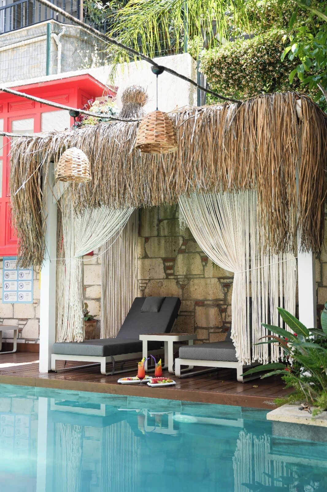 A poolside lounge area with a thatched canopy, two lounge chairs, and a stone wall backdrop.