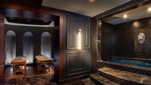 A spa area with a marble floor, wooden benches, a wall-mounted light, and a hexagonal tiled wall with a lion head sculpture. - TNR Otel & Spa (Kusadasi)