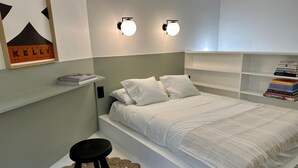 A bedroom with a bed, a small stool, a wall-mounted shelf with books, and two wall sconces. - Charming duplex apartment near Champs-Elysées (Paris)
