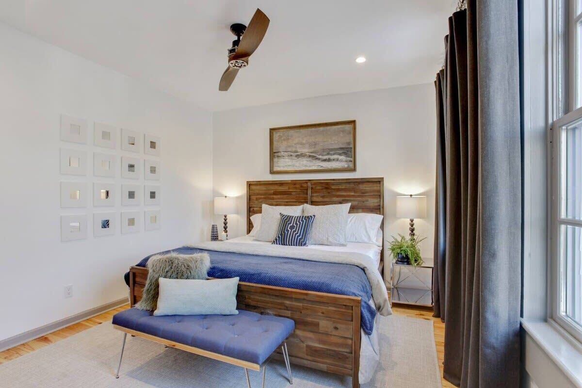 A bedroom with a wooden headboard, a bed with white bedding, a bench, a ceiling fan, and a large window with curtains.