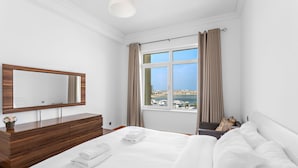 A hotel room with a large bed, a wooden dresser, a mirror, and a window with a view of a coastal city.
