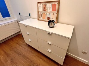 Una habitación con un tocador blanco, un collage enmarcado en la pared y un despertador negro encima.