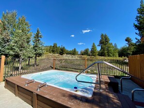 A hot tub with a wooden deck and metal railings, surrounded by a fence and trees.