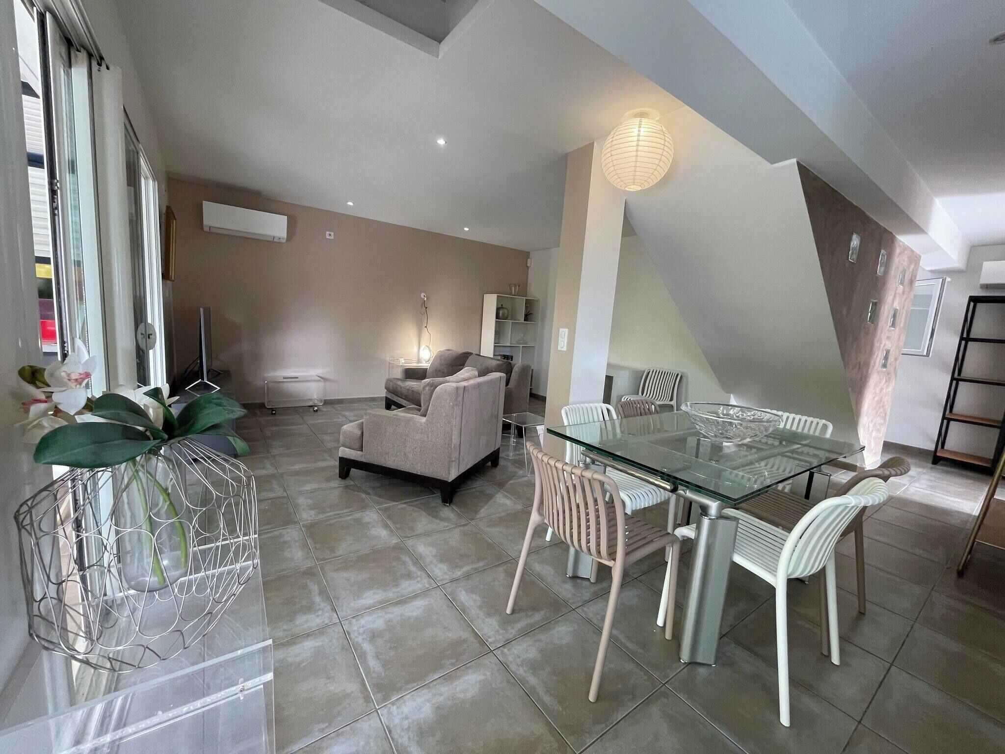 A modern living room with a glass dining table, grey sofas, and a large window.