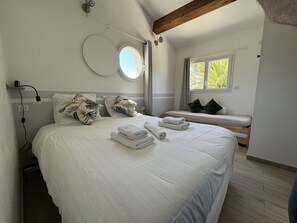 A bedroom with a large bed, a bedside table, a window with curtains, a round mirror, and a ceiling fan.