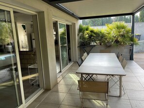 A covered patio area with a table and chairs, glass doors leading to a kitchen, and potted plants. - Sète, Jolie villa avec piscine à 5 mn des plages (Sète)