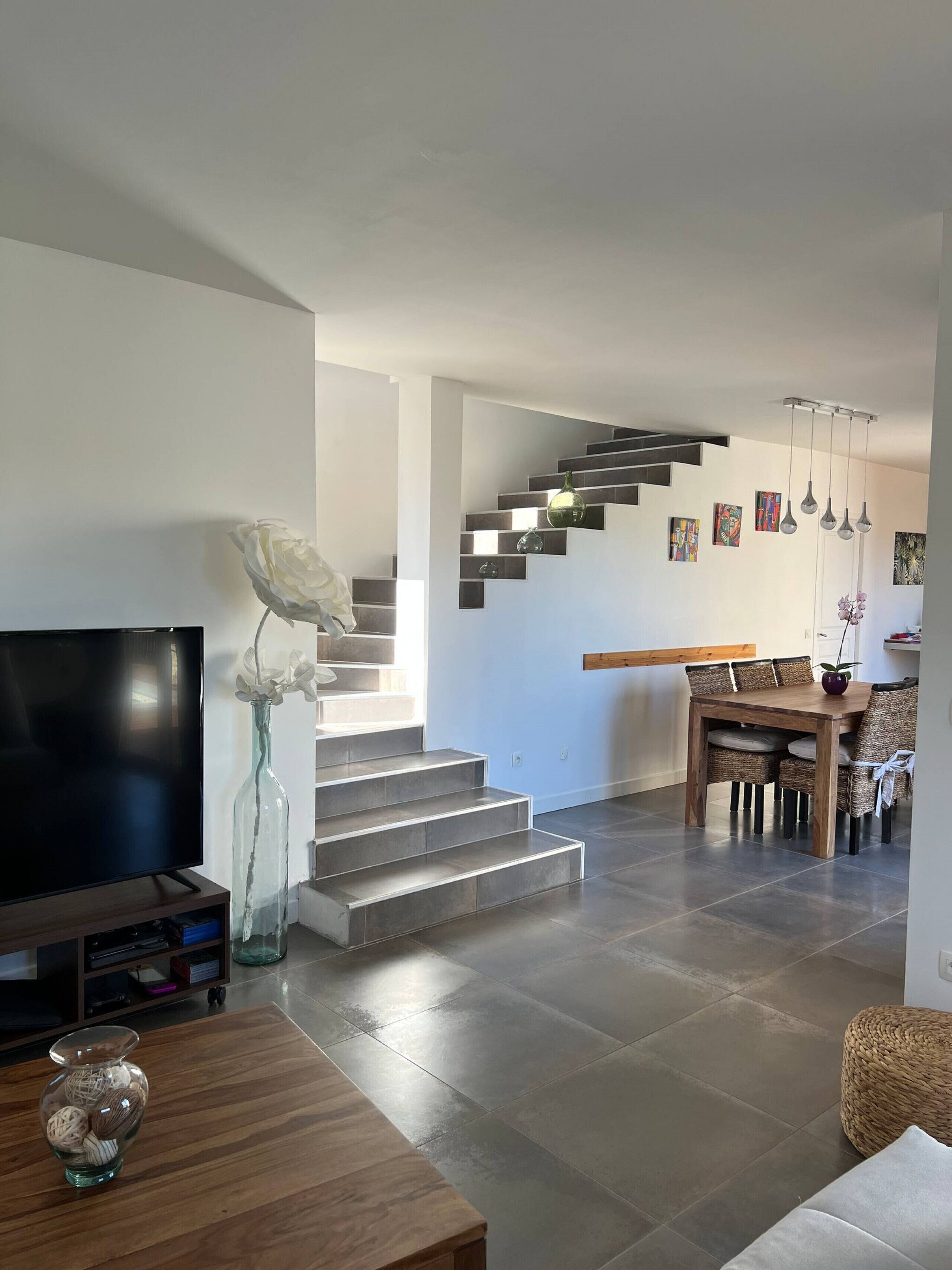 A modern living room with a flat-screen TV, a vase with white flowers, a wooden coffee table, and a staircase leading to a dining area with a wooden table and wicker chairs.