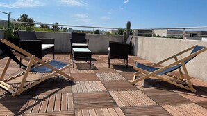 A rooftop patio with wooden flooring, two lounge chairs, a small table, and a cactus.