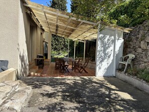 Een overdekt terras met een tafel en stoelen, een tuinhuisje en een stenen muur.