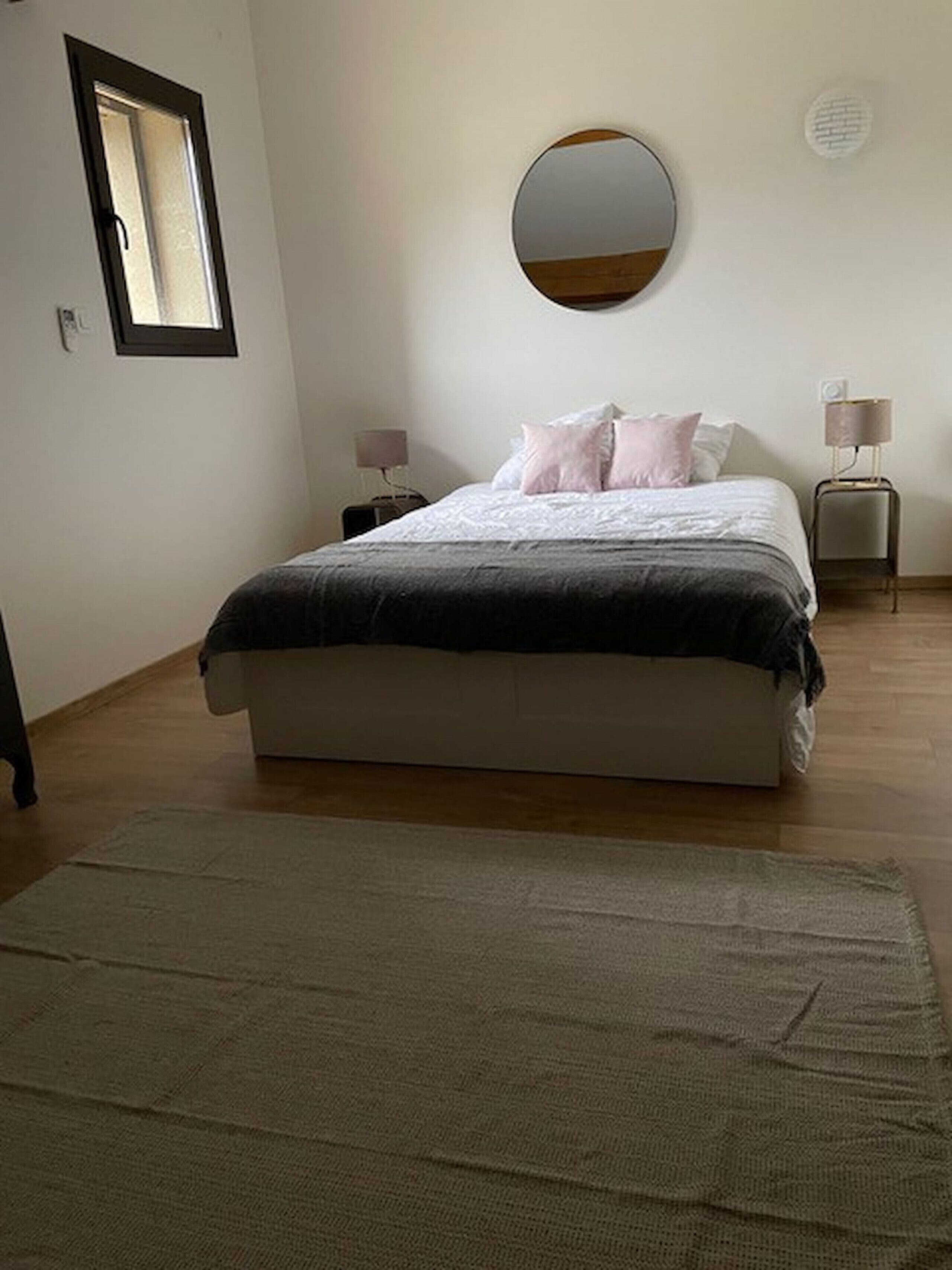 Une chambre à coucher avec un lit, un miroir rond accroché au mur, une table de chevet, une lampe et une fenêtre.