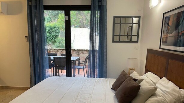 Una camera d'albergo con un letto grande, vista su un patio esterno attraverso una porta in vetro, un quadro appeso alla parete e un tavolino con sedie all'esterno.
