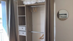 Een slaapkamer met een bed, een houten kledingkast met hangruimte, een klein krukje en een spiegel aan de muur.