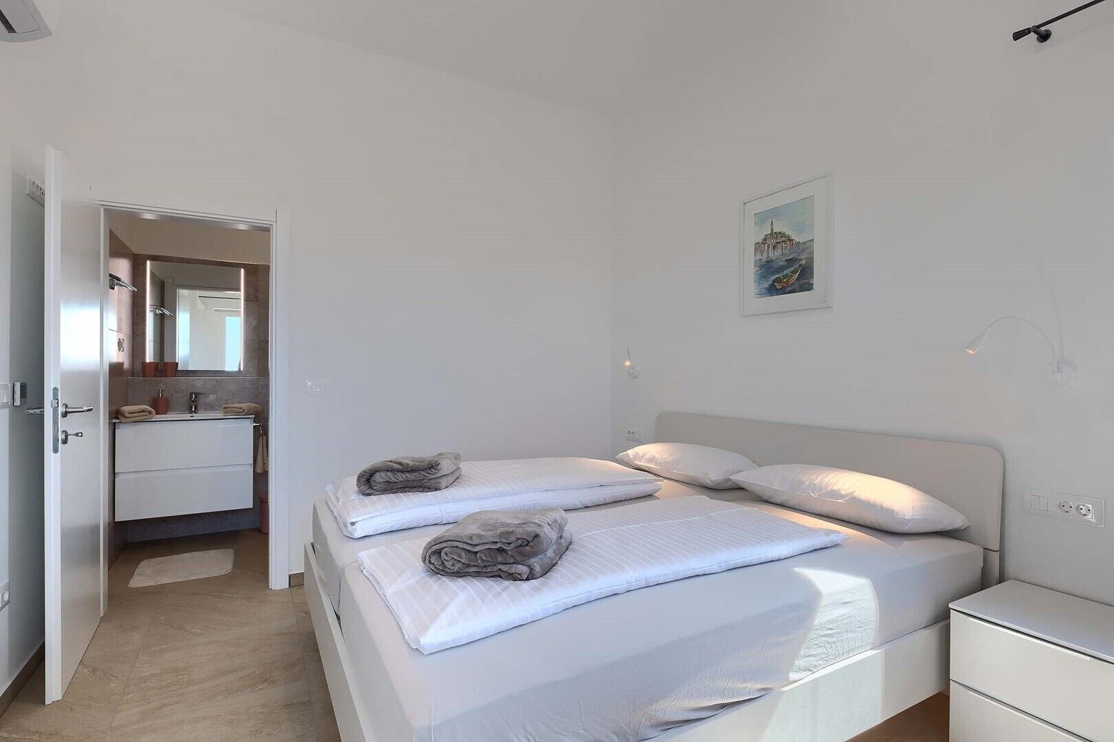 A bedroom with a bed, a bedside table, a nightstand, a bathroom with a sink and mirror, and a door leading to another room.