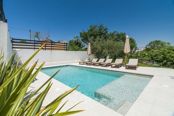 Une piscine rectangulaire à l’eau d’un bleu limpide, entourée d’un sol carrelé blanc. Six chaises longues sont alignées le long du bord de la piscine, chacune étant recouverte d’une housse beige. L’espace piscine est bordé d’un petit muret blanc et présente plusieurs plantes vertes au premier plan.